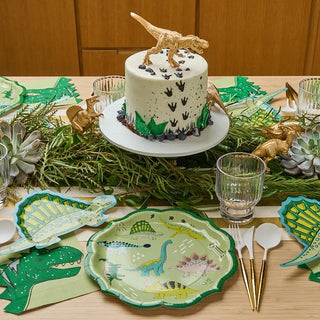 A Sophistiplate dinosaur-themed party table set with a white frosted cake topped with a golden t-rex, surrounded by greenery, elegant Dinosaur Dinner Plates, and matching decor.