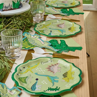 A festive table setting featuring Sophistiplate's whimsical Dinosaur Dinner Plates with ruffled green edges and matching napkins, complemented by elegant clear glasses and gold cutlery, all arranged on a natural wood table.