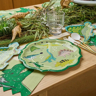 A festive table setting featuring elegant Sophistiplate Dinosaur Dinner Plates with ruffled green edges, golden cutlery, a clear glass, and a decorative greenery centerpiece with golden accents, on a wooden table.